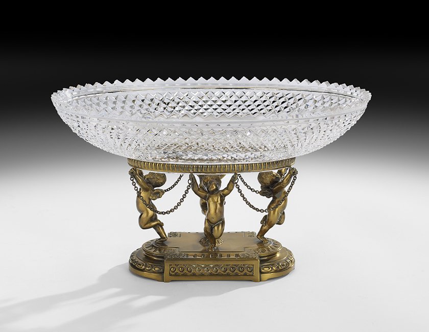 Regency-Style Glass and Brass-Plated Centerpiece: Regency-Style Cut Glass and Brass-Plated Centerpiece, mid-20th century, the brass-plated base decorated with cupids holding chains, around a pineapple finial, the bowl cut in the Waterford style, h. 7