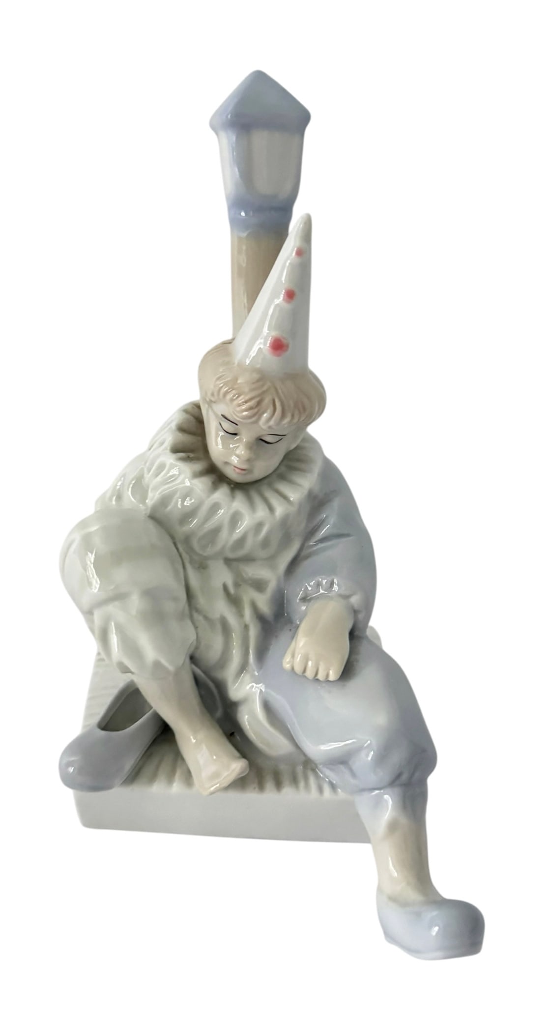 EUROPEAN PORCELAIN SEATED PIERROT CLOWN FIGURINE: Vintage glazed porcelain figurine depicting a seated Pierrot-style clown wearing a pointed hat and ruffled collar, modeled beside a tall street lamp. Decorated primarily in soft white and pale blue-gr