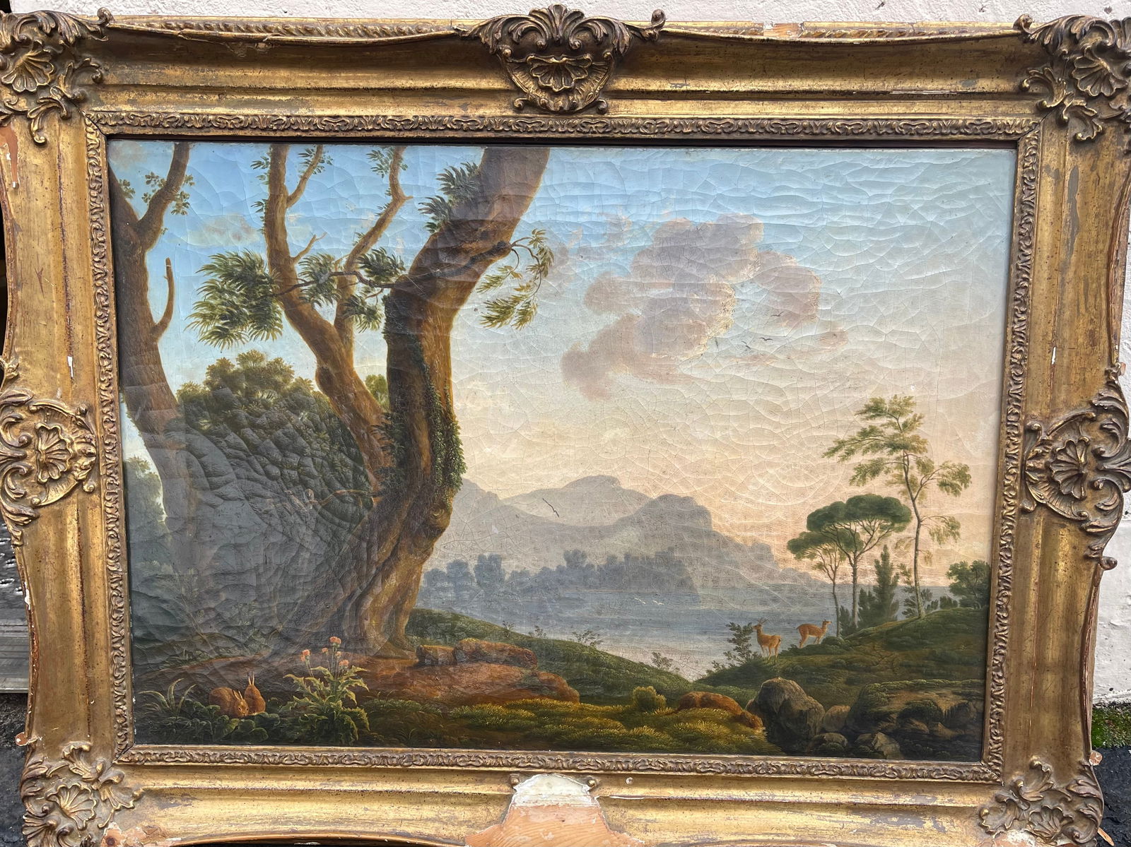 Landscape Painting Italian School 19th Century: An Italian school painting of a landscape with a large tree on the left side with three rabbits near a tree trunk. A Lake is in the mid ground with mountains with a blue sky with wispy cloud in the ba