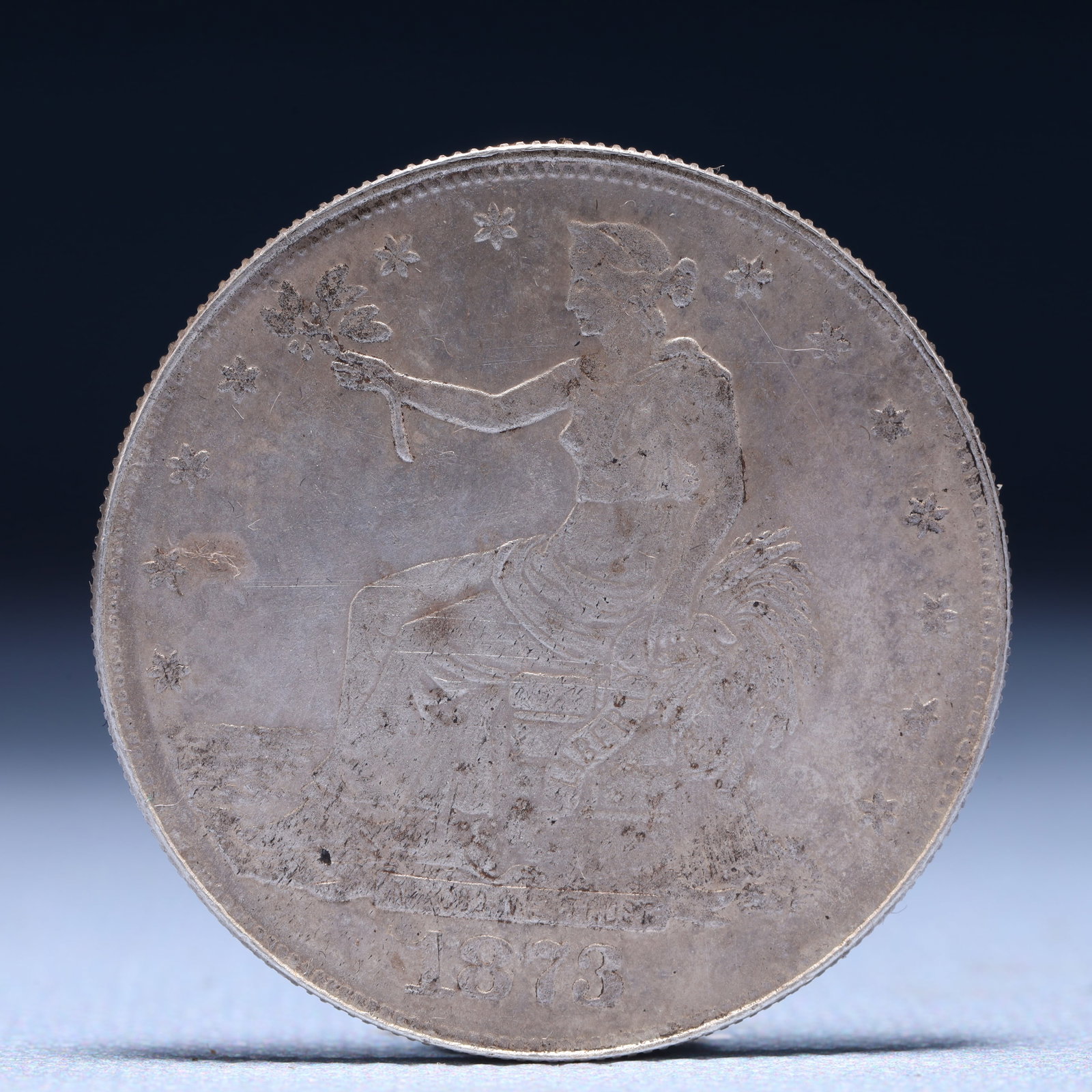 USA Trade Dollar 1873 Coin: American coin. Obverse: Liberty seated on a pedestal facing the sea to the left holding olive branches surrounded by 13 stars. IN GOD WE TRUST / LIBERTY / 1873. Reverse: An eagle holding branches with