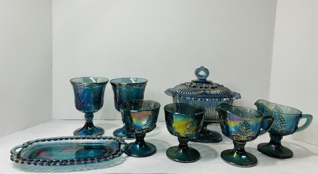 A Collection of Blue Carnival Glass Tableware in the Grape and Leaf Pattern: A collection of blue iridescent carnival glass tableware featuring a molded grape and leaf design, resembling the Harvest Grape pattern produced by Indiana Glass. The group includes multiple footed go