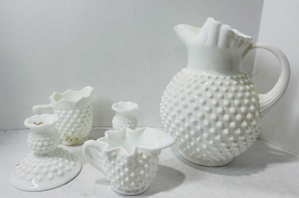 A Collection of Fenton White Milk Glass Hobnail Tableware: A collection of Fenton white milk glass items in the hobnail pattern. The assortment includes a large bulbous pitcher featuring a ruffled rim and an applied ribbed handle. Additional pieces include mu