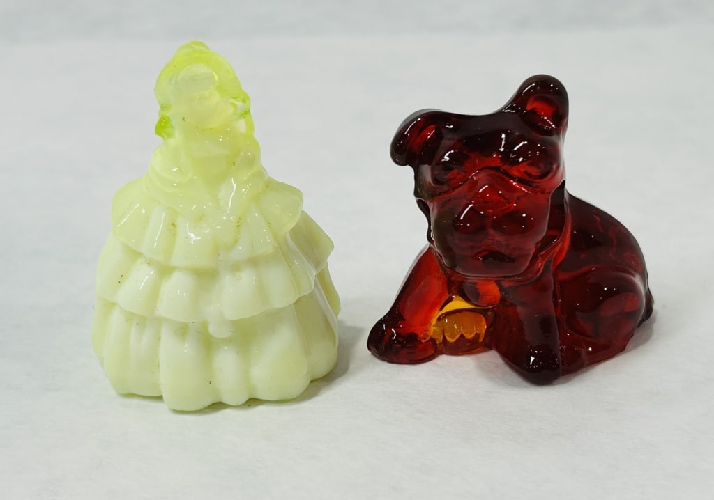 Two Vintage Pressed Glass Figurines including a Uranium Custard Glass Lady and an Amberina Bulldog: This collection features two vintage pressed glass miniatures. The first is a lady in a tiered crinoline dress, rendered in opaque yellow custard glass. This piece exhibits strong green fluorescence u