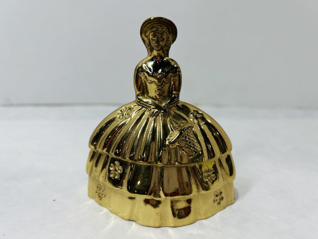 Vintage Brass Figural Table Bell in the Form of a Southern Belle: This vintage table bell is cast from a gold-toned metal, likely brass, in the form of a female figure dressed in mid-19th-century style attire. The figure features a wide-brimmed sun hat and a three-t