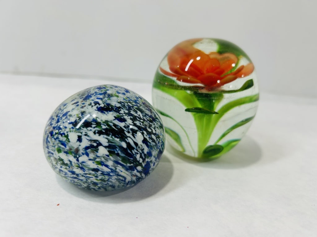 Two Art Glass Paperweights with Lampworked and Frit Inclusions: This lot features two distinct glass paperweights. The first is a clear glass spherical weight containing an internal lampworked orange flower with multiple tiers of petals and green stems and foliage