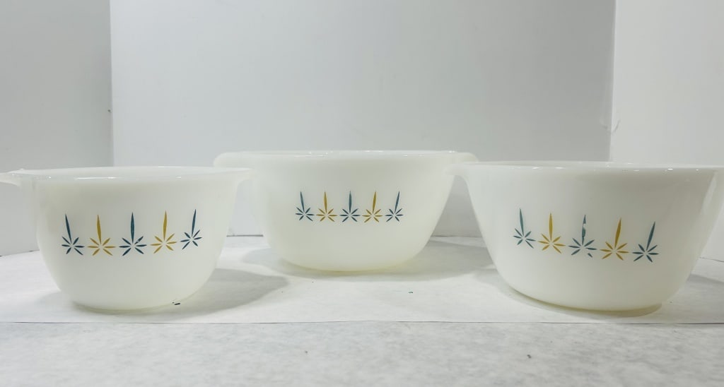 White Milk Glass Nesting Bowls with Mid-Century Starburst Motif: A set of three nesting bowls constructed from opaque white milk glass. Each bowl features a flared rim with integrated tab handles located on opposite sides. The exterior of each vessel is decorated w