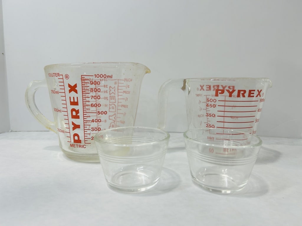 A Group of Vintage Pyrex Glass Measuring Cups and Custard Cups: A group of vintage clear glass kitchenware manufactured by Pyrex. The assortment consists of two graduated measuring cups and two small custard cups. The measuring cups feature red printed graphics in