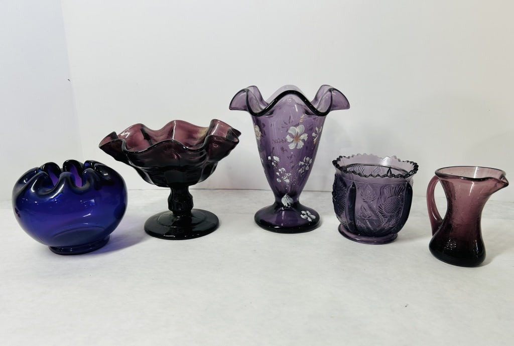 A Collection of Amethyst and Cobalt Decorative Glass Items Including Fenton Art Glass: A collection of assorted decorative glass items, featuring pieces by the Fenton Art Glass Company. The group primarily consists of amethyst and cobalt blue glass and includes a pedestal compote with a
