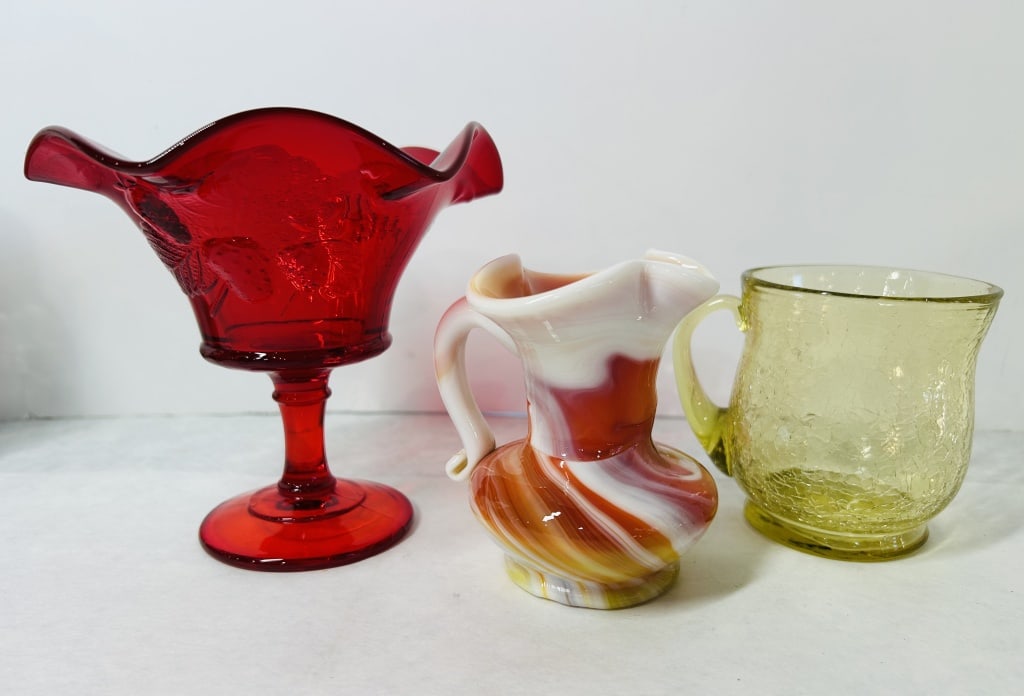A Trio of Vintage Decorative Glassware Including a Ruby Red Compote, Slag Glass Pitcher, and Yellow: This collection consists of three individual pieces of decorative glassware. The first piece is a ruby red pressed glass compote featuring an embossed strawberry and foliage pattern. The vessel is des