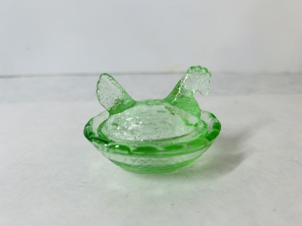 A Miniature Green Pressed Glass Hen-on-Nest Salt Cellar: This miniature two-piece pressed glass covered dish is designed in the form of a hen on a nest, a traditional style often used as a salt cellar. The dish is made of translucent green glass. The figura
