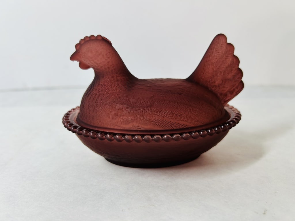 Satin Burgundy Glass Hen on Nest Covered Dish: The glass is a deep burgundy or plum red color, displaying translucent amethyst or ruby tones in thinner areas when viewed against the light. The lid is molded into the form of a nesting hen with deta