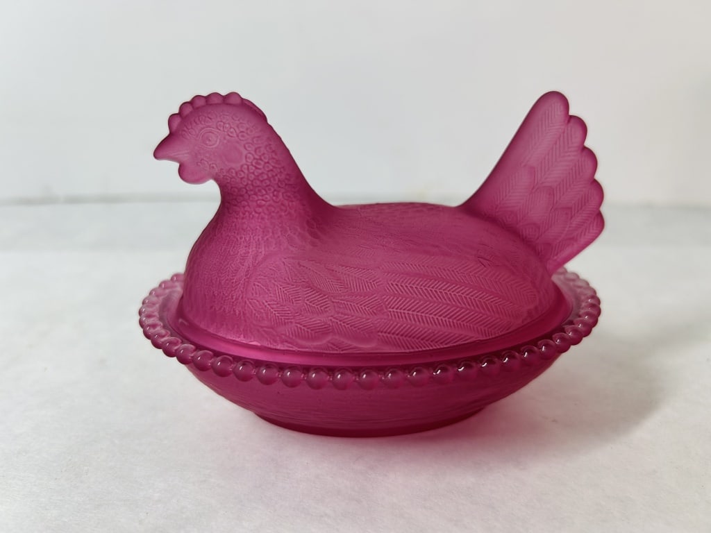 Satin Fuchsia Glass Hen on Nest Covered Dish: This two-piece novelty dish is crafted in the form of a nesting hen. The set consists of a lid and a base, both made from pressed glass with a uniform satin finish. The glass is a vibrant, deep fuchsi
