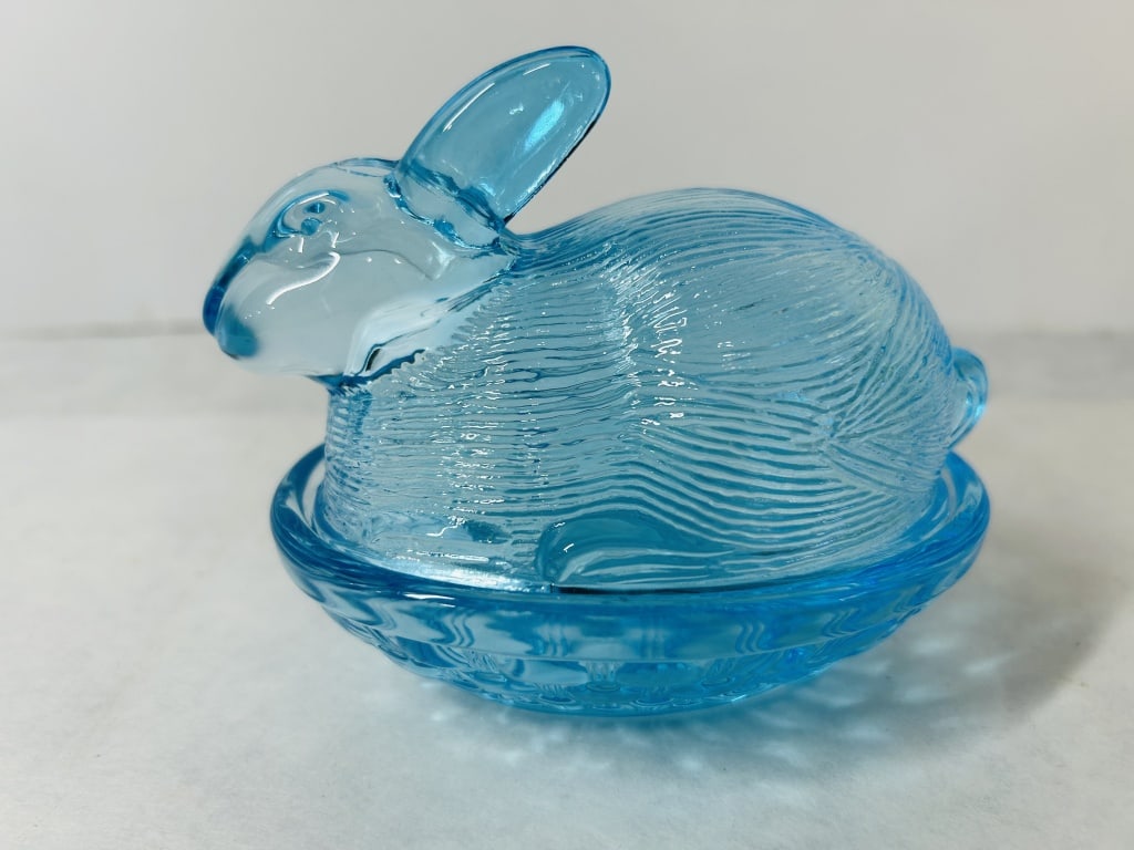 Blue Pressed Glass Bunny on Basket Covered Dish: This two-piece decorative covered dish is crafted from translucent light blue pressed glass, a form commonly known as a Rabbit on Nest or Bunny on Basket. The set consists of a lid in the shape of a s
