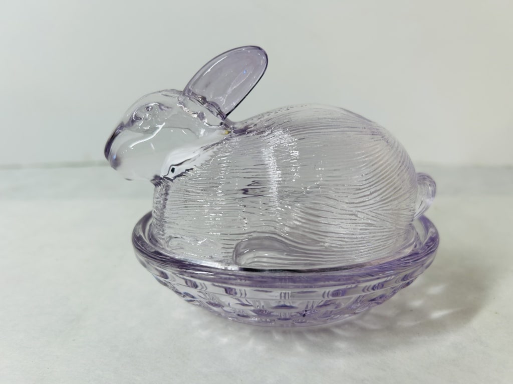 Lavender Pressed Glass Rabbit on Nest Covered Candy Dish: This two-piece pressed glass covered dish is molded in the form of a rabbit resting on a nest. The glass is translucent with a pale lavender or light amethyst tint. The lid is shaped as a crouching ra