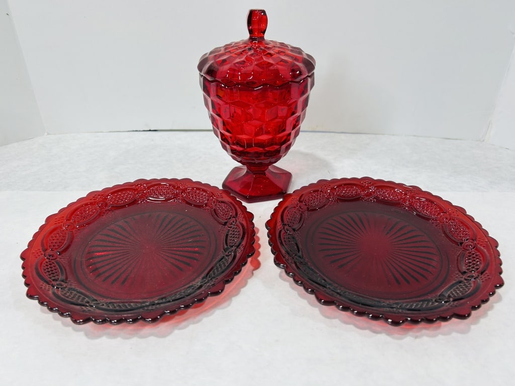 A Trio of Ruby Red Pressed Glass Tableware in the 1876 Cape Cod Pattern: This three-piece set of ruby red pressed glassware is in the 1876 Cape Cod pattern, a design produced for the Avon collection beginning in the 1970s. The set consists of two plates and one lidded pede
