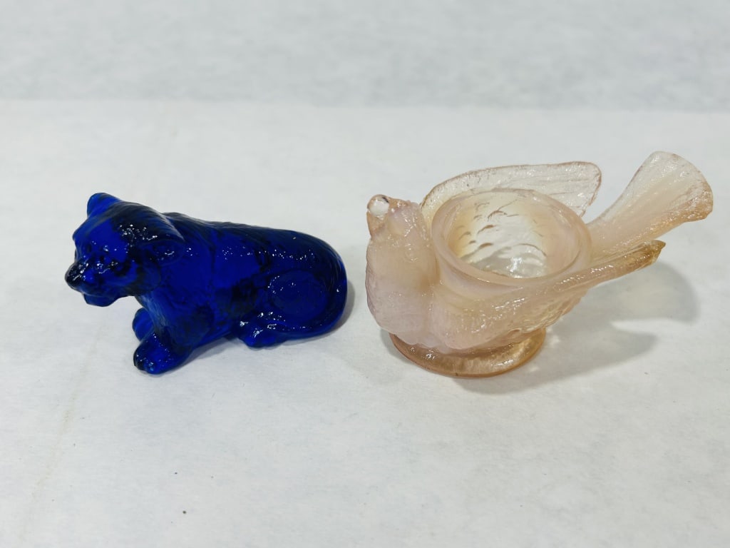 Two Decorative Glass Items: Cobalt Blue Cat Figurine and Pink Opalescent Bird Salt Cellar: This lot features two decorative glass objects. The first is a figurine depicting a reclining big cat, such as a mountain lion or cougar, rendered in translucent cobalt blue pressed glass. The second 