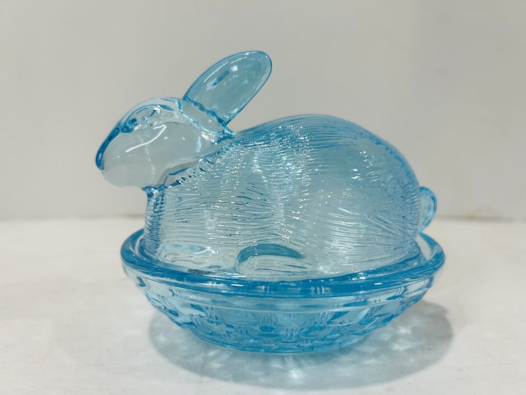 Transparent Blue Pressed Glass Rabbit on Nest Covered Dish: This item is a two-piece pressed glass covered dish featuring a lid molded in the form of a crouching rabbit resting on a basket-weave base. The piece is crafted from transparent light blue glass, a s