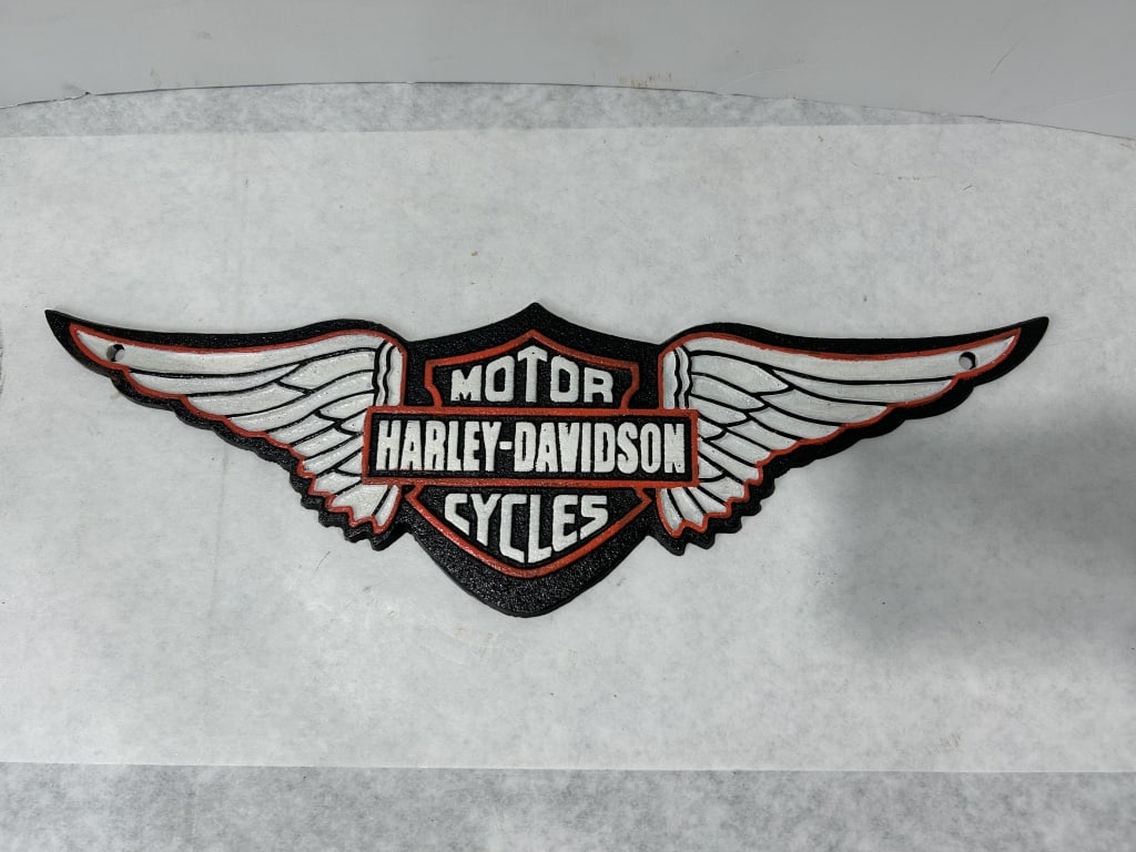 Harley-Davidson Winged Bar and Shield Logo Wall Plaque: A cast metal wall plaque featuring the classic Harley-Davidson Bar and Shield logo flanked by stylized eagle wings. The piece is constructed with a raised relief design set against a pebbled black bac