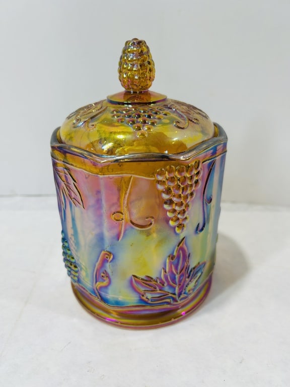 Marigold Carnival Glass Covered Jar in the Harvest Grape Pattern: This marigold carnival glass covered jar is produced in the Harvest Grape pattern, attributed to the Indiana Glass Company. The item, likely intended as a canister or biscuit jar, is made of pressed g