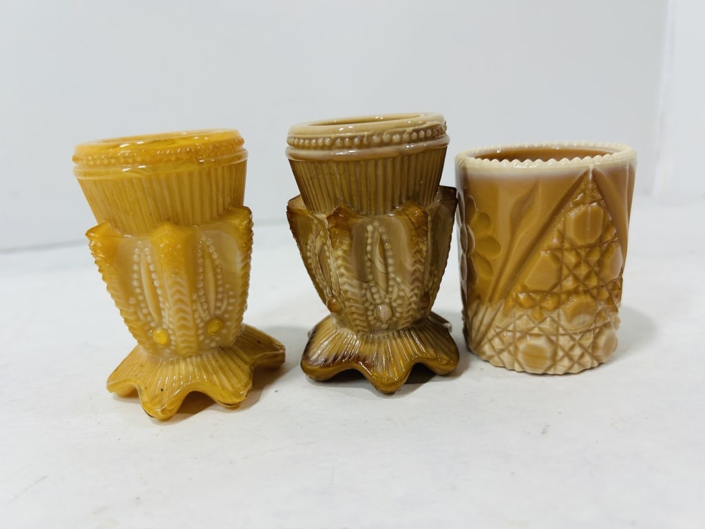 A Trio of Caramel Slag Glass Toothpick Holders in Thistle and Geometric Patterns: A collection of three small pressed glass vessels crafted in caramel slag glass, an opaque marble glass also known as chocolate glass. Two of the vessels are footed toothpick holders featuring the Thi