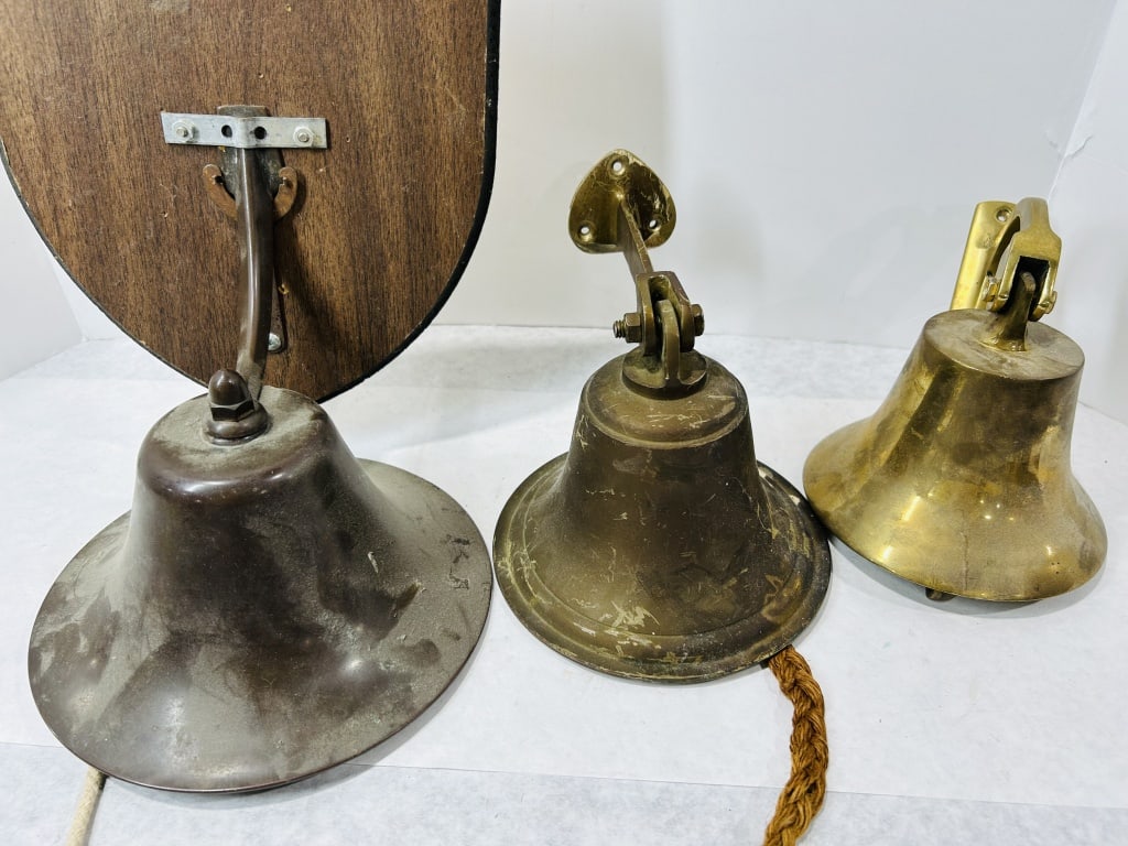 A Trio of Vintage Brass and Bronze Nautical Ship's Bells: This lot consists of a trio of vintage nautical-style wall-mounted bells of varying sizes and designs. The largest bell features a dark brown patinated finish, likely bronze or treated brass, and is m