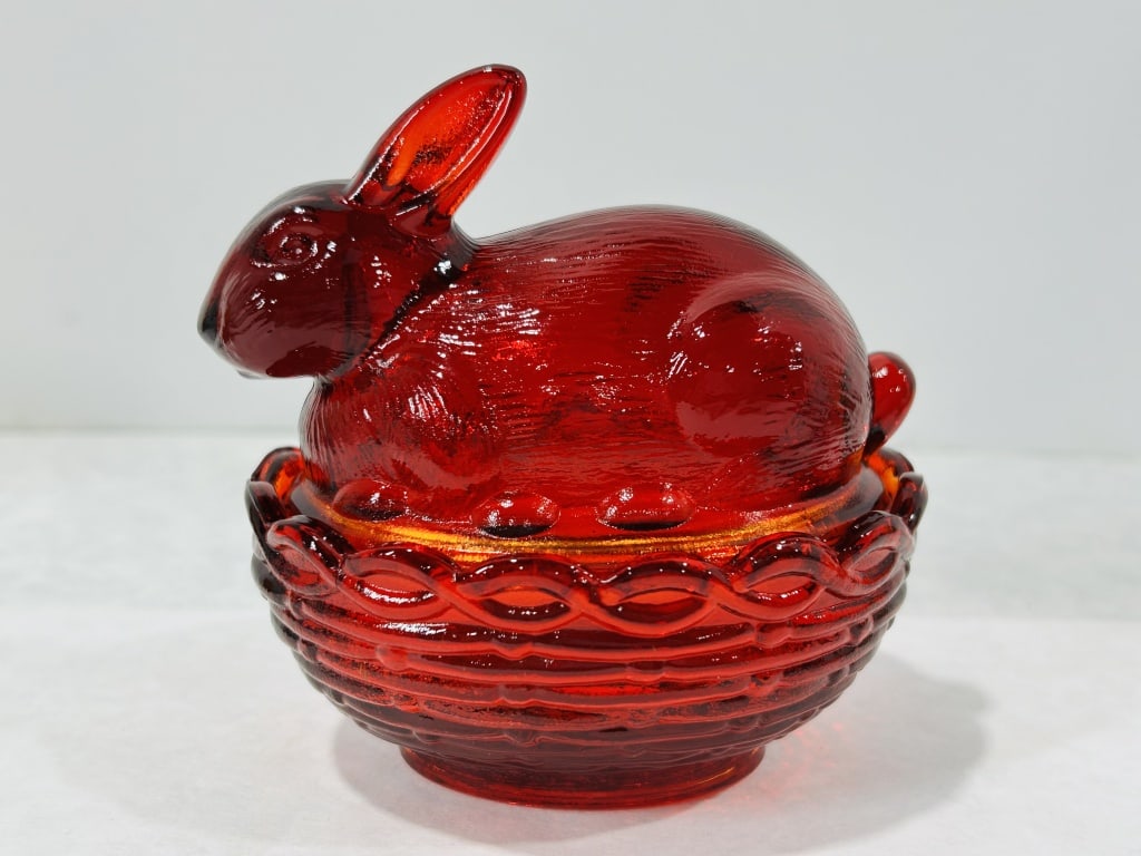 Ruby Red Pressed Glass Rabbit on Nest Covered Dish Mosser Glass: This two-piece covered dish in ruby red pressed glass is attributed to Mosser Glass. The design features a rabbit lid resting on a basket-weave base. The lid is detailed with textured fur, alert ears,