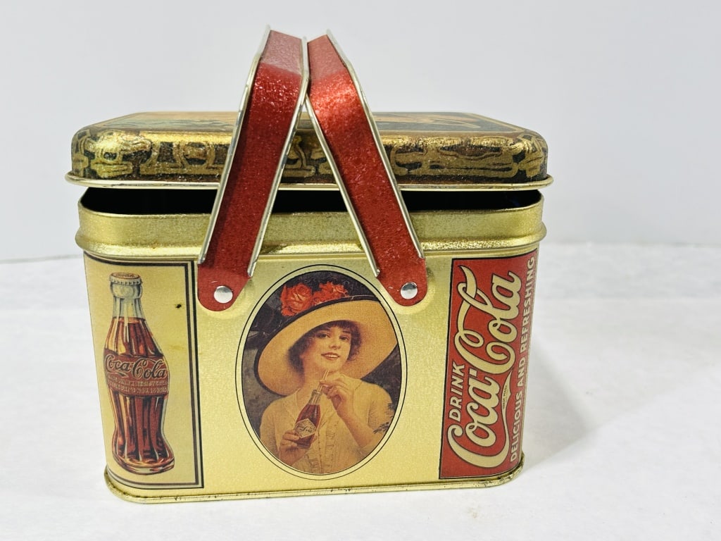 Promotional Coca-Cola Lithographed Tin Picnic Basket with Dual Swivel Handles: This promotional Coca-Cola metal storage tin is designed in the form of a small picnic basket. It features a removable lid and two red-coated metal swivel handles secured with rivets. The exterior sur