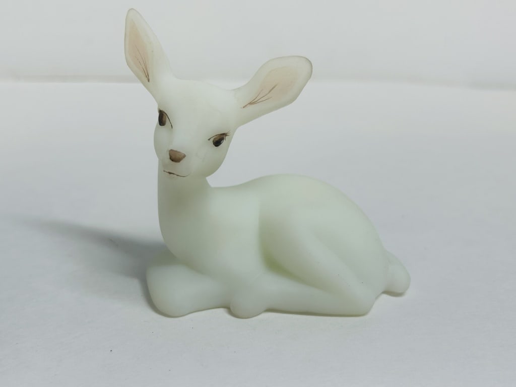 FENTON WHITE SATIN GLASS FAWN: A reclining white milk glass fawn figurine with a satin finish produced by the Fenton Art Glass Company. The hand-painted piece features the signature of decorator K. Caves and retains the original ma