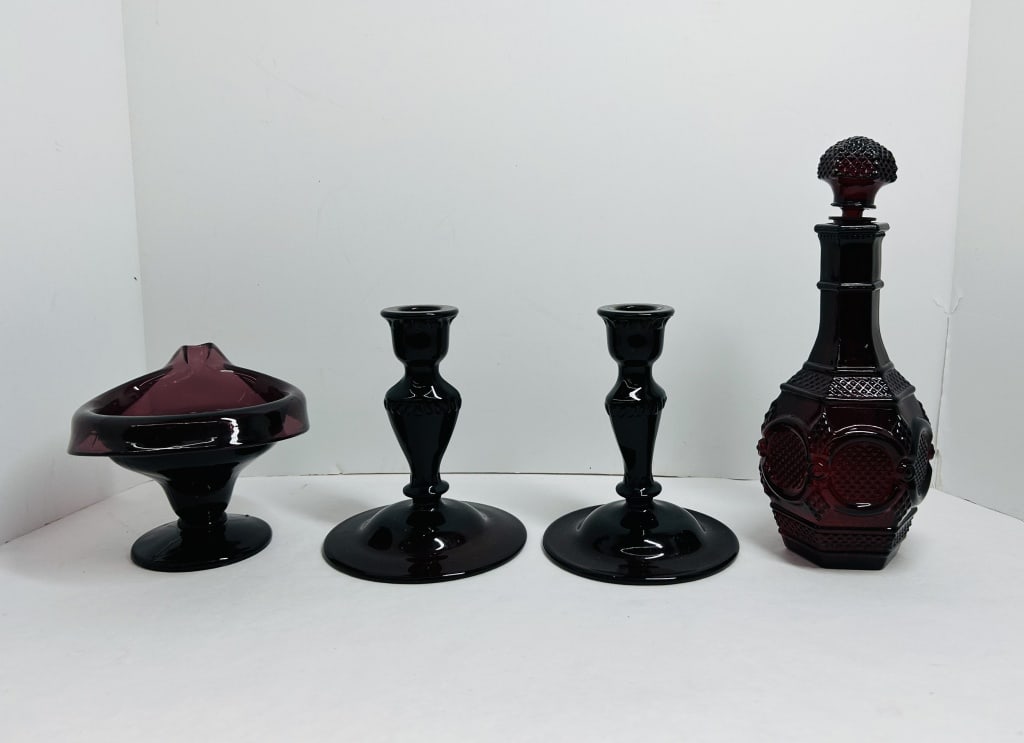AVON AND AMETHYST GLASS, 1970S: Avon ruby red decanter in the 1876 Cape Cod pattern and coordinating amethyst glass pieces. The collection includes a pair of black amethyst glass candlesticks and a footed amethyst glass bowl with a 