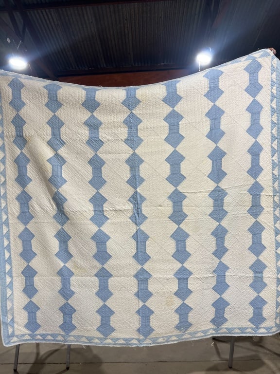 BLUE AND WHITE COTTON SPOOL QUILT, MID-20TH CENTURY: Mid-20th-century hand-quilted cotton patchwork quilt featuring a two-color Spool pattern with a sawtooth border. The reverse consists of solid white cotton fabric displaying dense hand-quilted
