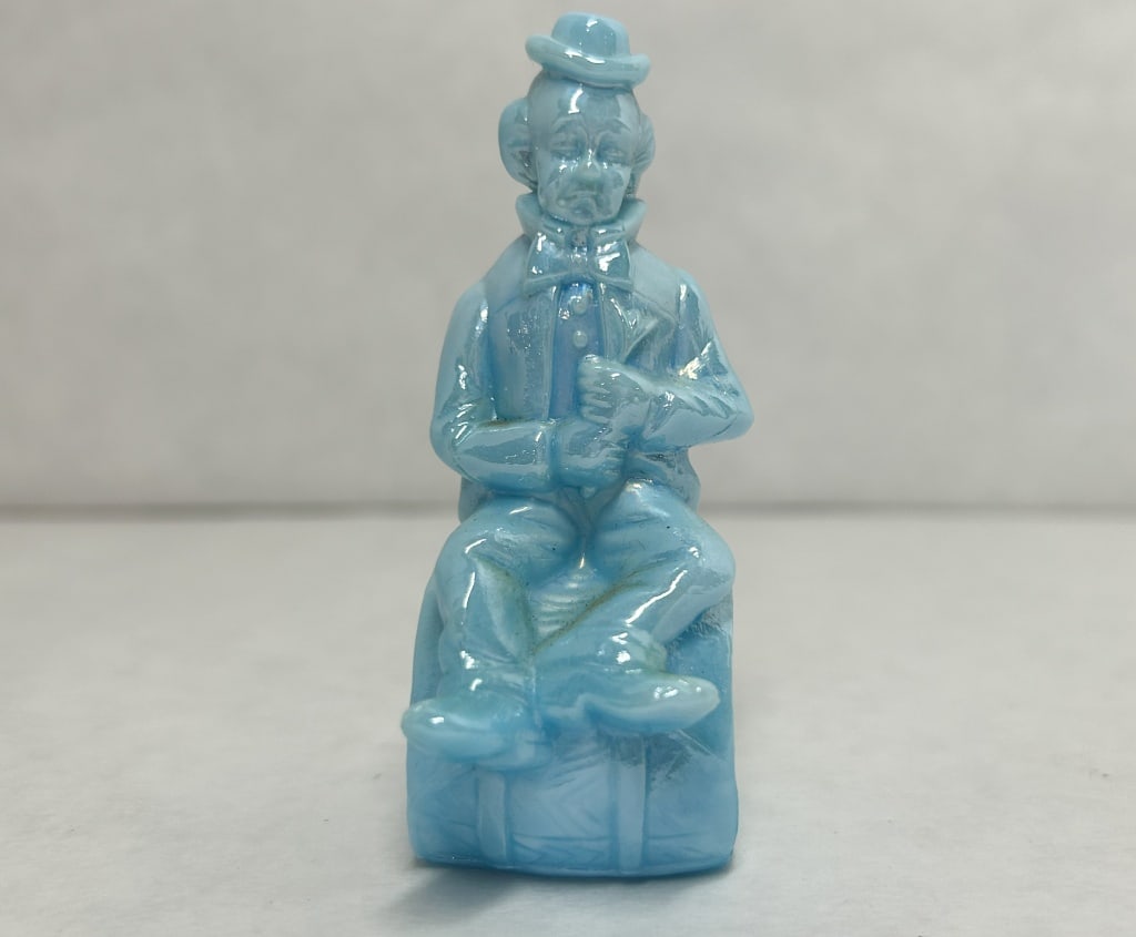 MOSSER BLUE SLAG CLOWN FIGURINE 1982: A blue iridescent slag glass figurine representing the character Poko, produced by the Mosser Glass Company. One end of the barrel base is molded with a stylized letter M over the year 1982, and the r