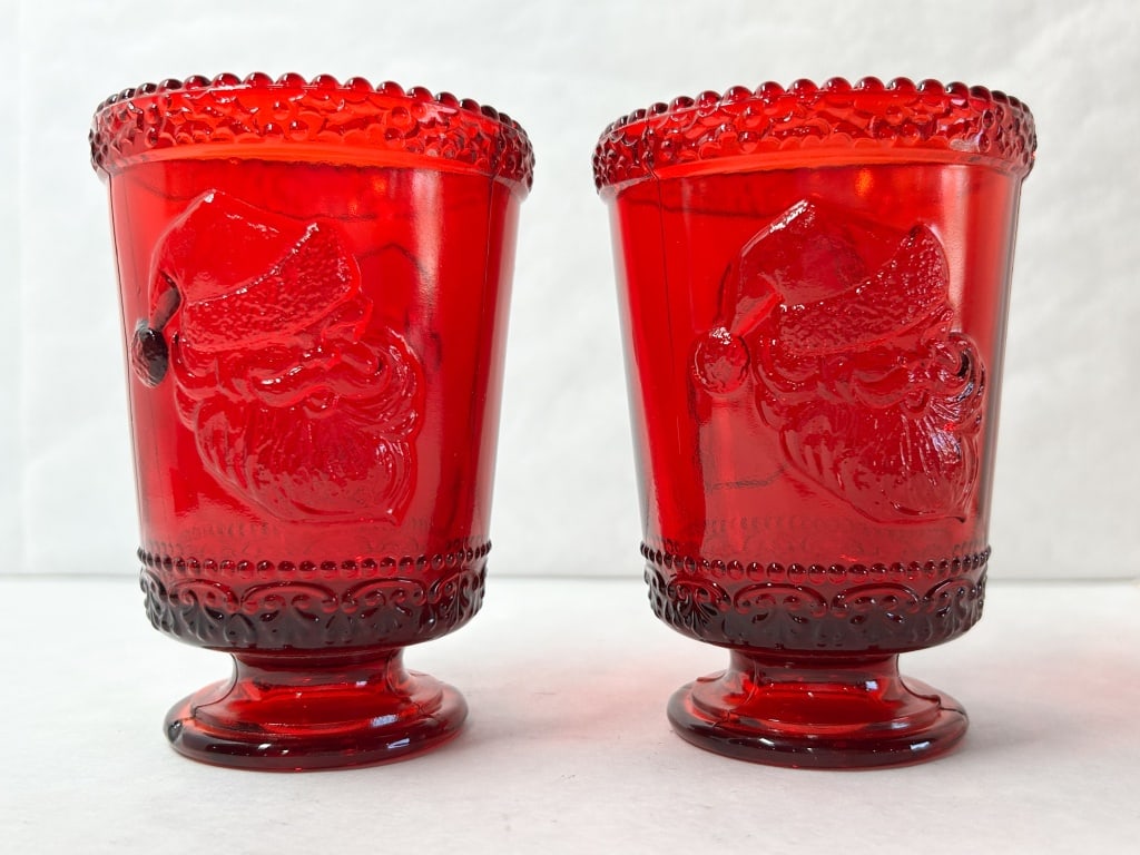 MOSSER RUBY RED GLASS SANTA CUPS: A pair of ruby red pressed glass Santa cups featuring dual embossed portraits and beaded rims. Each base bears an impressed monogram M within a beveled square frame.