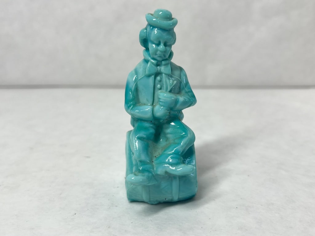 MOSSER BLUE SLAG PERFORMER GLASS CLOWN, 1982: A turquoise blue slag glass clown figurine produced by Mosser Glass. The character is modeled seated on a barrel embossed with the letter M above the year 1982 on one end and THE END on the other.