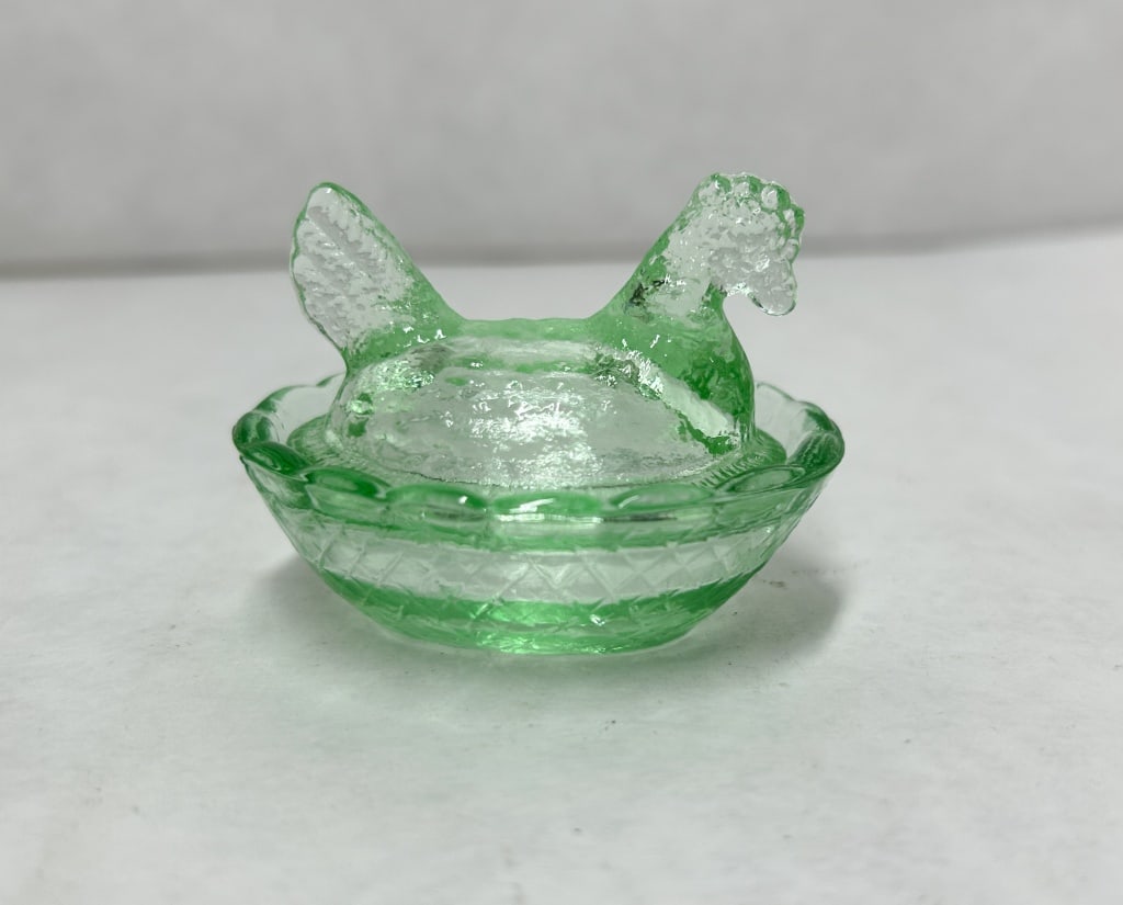 MINIATURE GREEN GLASS HEN ON NEST: A two-piece miniature pressed glass covered dish in transparent light green, modeled as a hen seated upon a basketweave nest. The lid features stippled feather detailing and a prominent comb, while th
