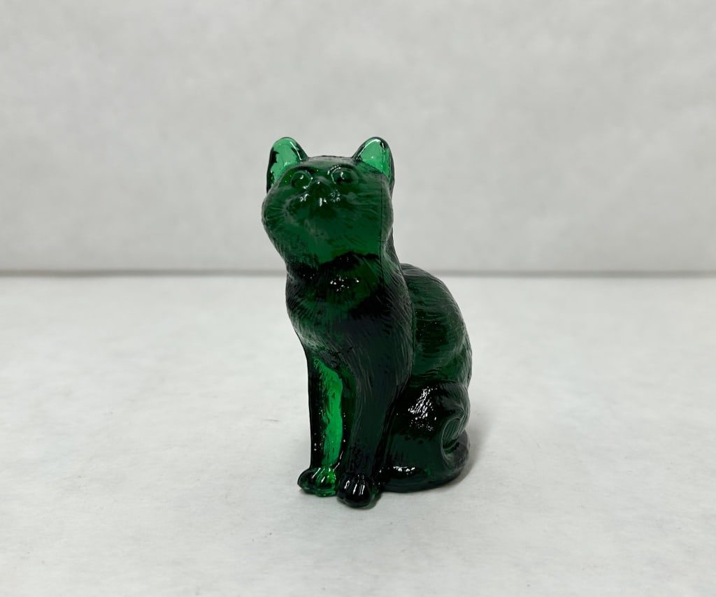 MOSSER EMERALD GREEN GLASS SITTING CAT: Seated cat figurine crafted from transparent emerald green glass. The piece features a molded surface texture simulating fur and the head is posed tilted upward.