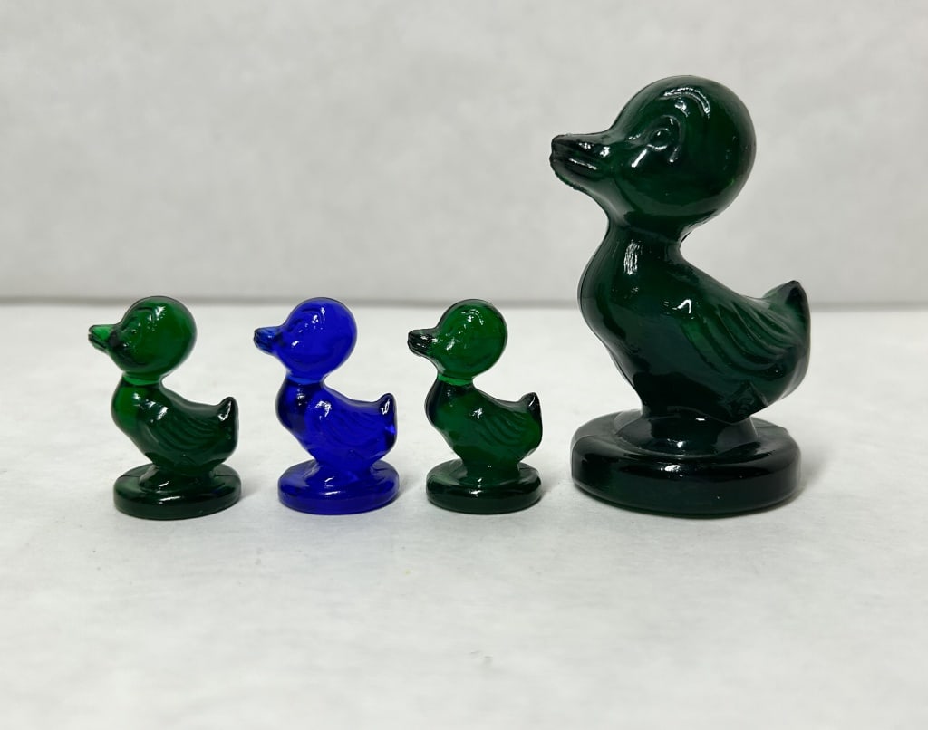 BOYD DEBBIE DUCK GREEN: A set of four pressed glass figurines including one large duck and three smaller ducklings in forest green and cobalt blue. An embossed diamond mark containing the superimposed letters I and G is loca