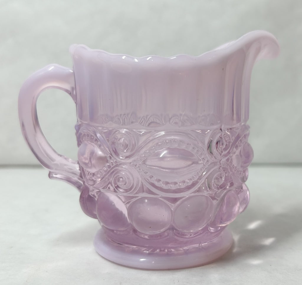 MOSSER PINK SLAG GLASS EYEWINKER CREAMER: Pressed slag glass creamer in the Eyewinker pattern featuring marbled pink and white coloration. The mold-formed vessel displays almond motifs and scrolls beneath a scalloped rim.