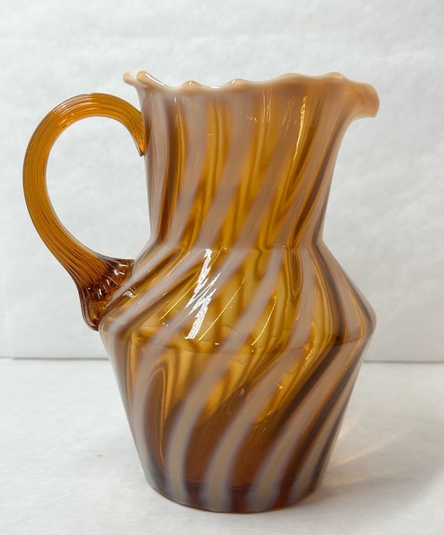AMBER OPALESCENT SPIRAL GLASS PITCHER: An amber opalescent glass pitcher featuring a diagonal spiral pattern and a ruffled rim. The vessel is fitted with an applied ribbed handle with a multi-pronged terminal.