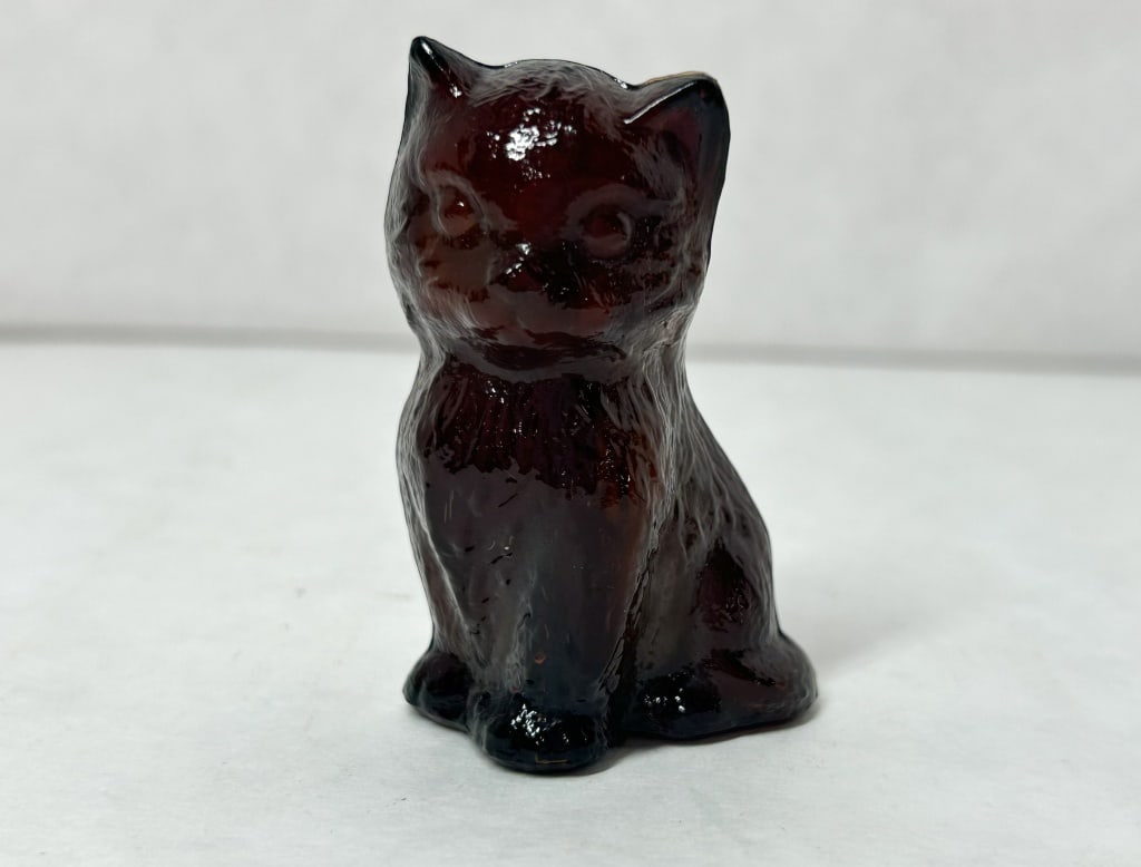 BOYD MISS COTTON DARK AMBER GLASS KITTEN: A small figurine of a seated kitten crafted from pressed dark amber glass, appearing opaque black under standard lighting with deep reddish-brown translucency visible at the ears.