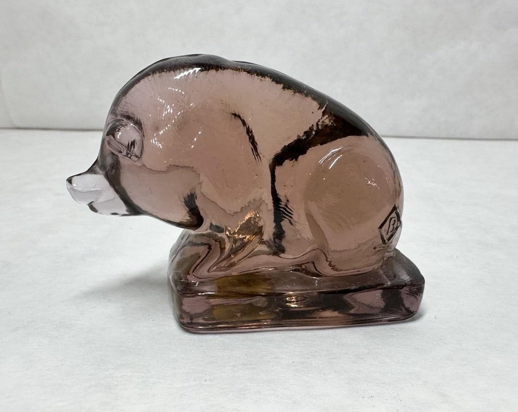 BOYD AMETHYST SUEE GLASS PIG: A pressed glass figurine of a crouching pig or boar, crafted from transparent amethyst glass and mounted on a solid rectangular base. The piece features a vertical mold seam along the center and a rai