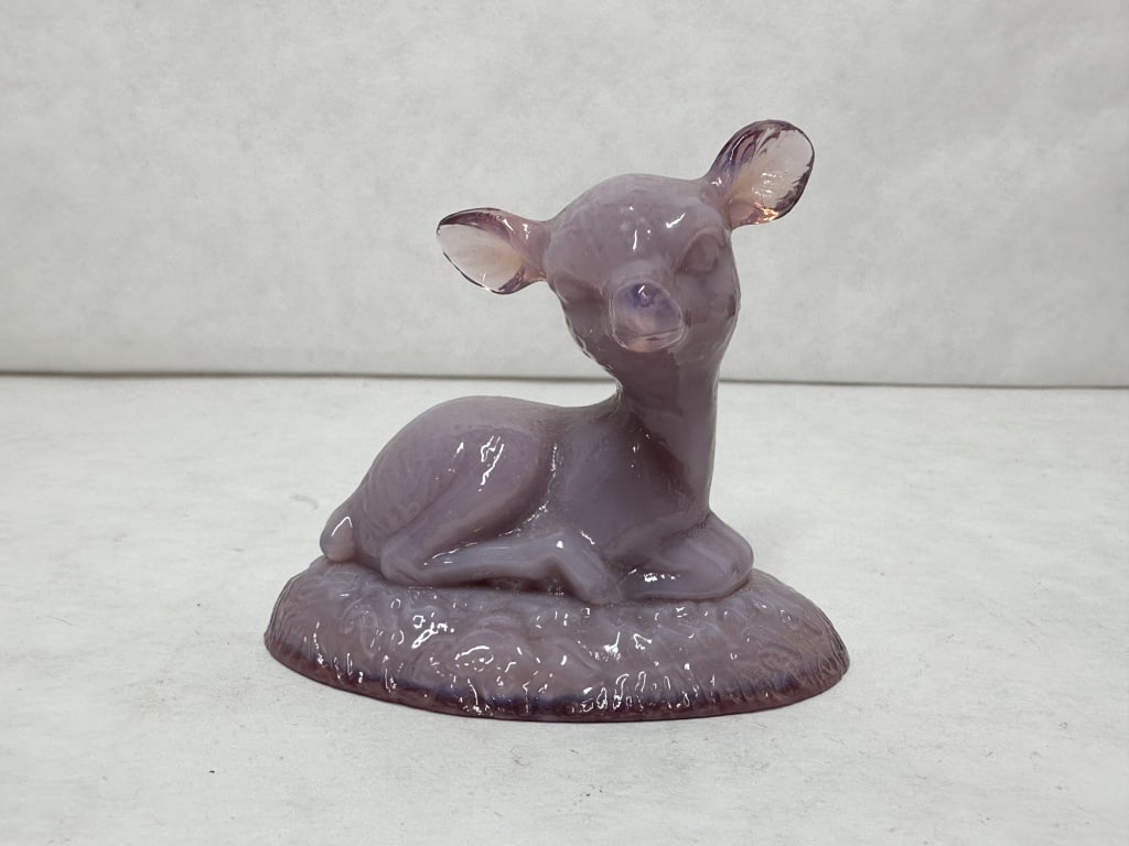 BOYD BINGO DEER PURPLE GLASS: A purple slag glass figurine depicting a recumbent fawn with prominent perked ears. The figure rests on a textured oval base and displays characteristic lavender and purple marbling throughout the