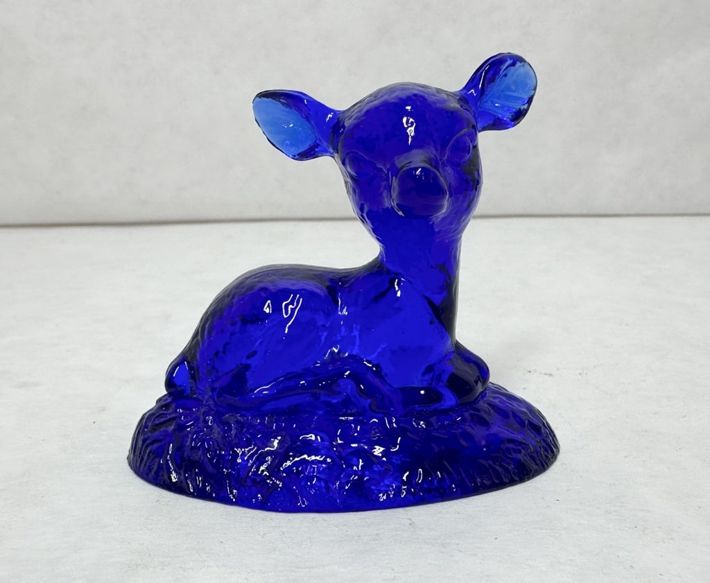 BOYD BINGO DEER COBALT BLUE GLASS, 1970S: A pressed glass figurine of a reclining fawn rendered in deep cobalt blue. The figure is posed on an oval, textured base with a high-gloss surface finish.