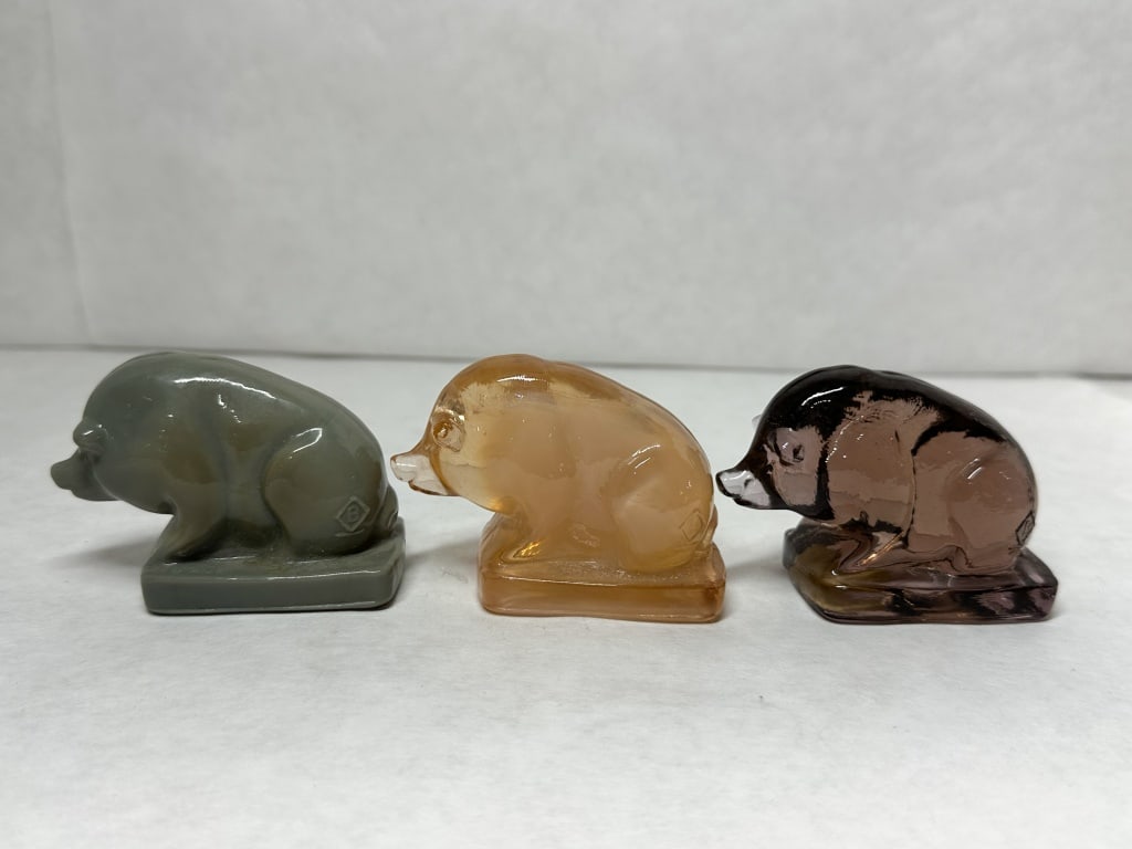 BOYD SUEE THE PIG GLASS: A trio of stylized pressed glass pig figurines produced by Boyd's Crystal Art Glass in opaque grey-green slag, amber, and black amethyst glass. Each example is hunched on a rectangular base and featur