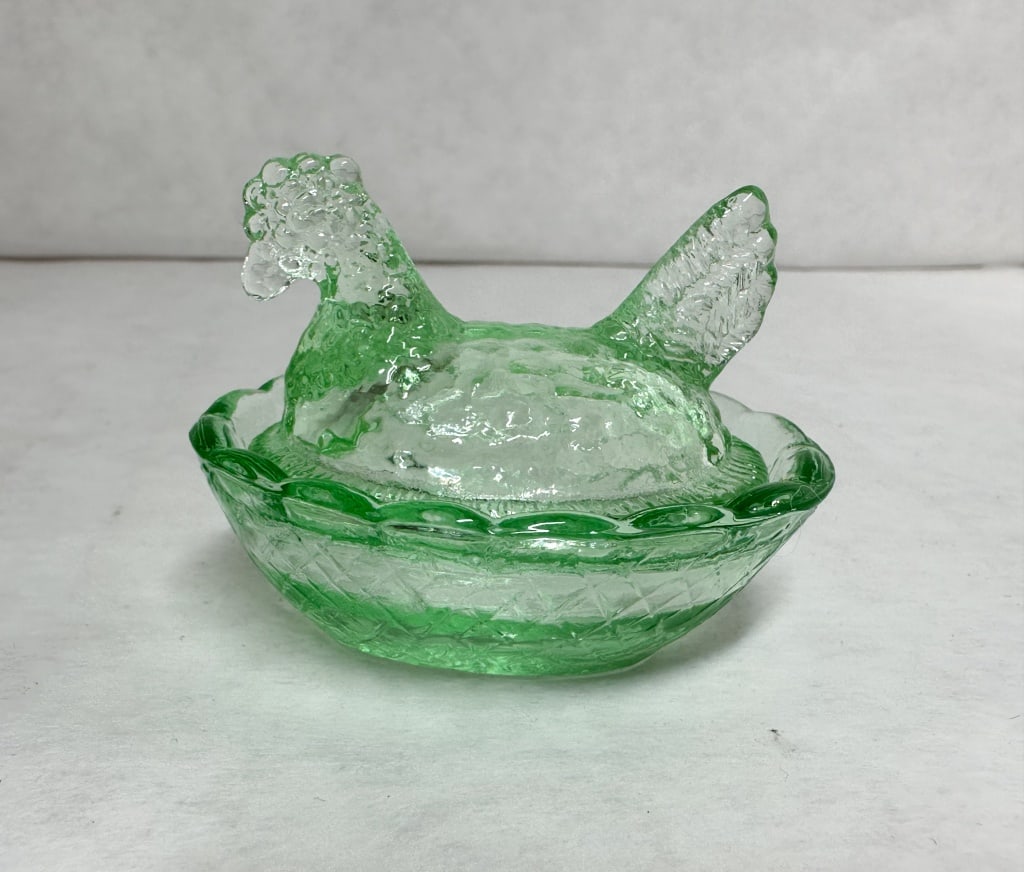 GREEN PRESSED GLASS HEN ON NEST: Vintage two-piece covered dish in the form of a nesting hen, crafted from translucent green pressed glass. The hen lid features a stippled texture and feathered tail, resting on a base with a basketwe