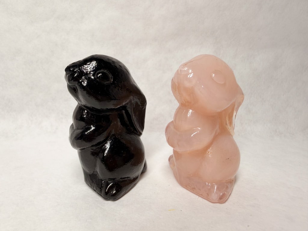 BOYD GLASS RABBIT FIGURINES, 1989-1993: A pair of pressed glass rabbit figurines produced by Boyd's Crystal Art Glass in opaque black and pink slag glass. Each figurine bears a raised mark on the lower side consisting of a letter B within a