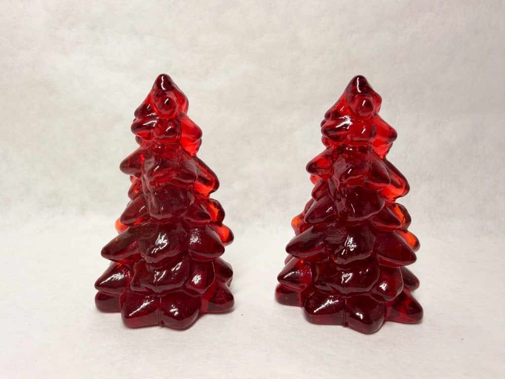 MOSSER MINI RUBY GLASS TREES: A pair of stylized Christmas tree figurines crafted from molded ruby red glass. The forms feature rounded, tiered branches tapering to a peak with a glossy, translucent finish.