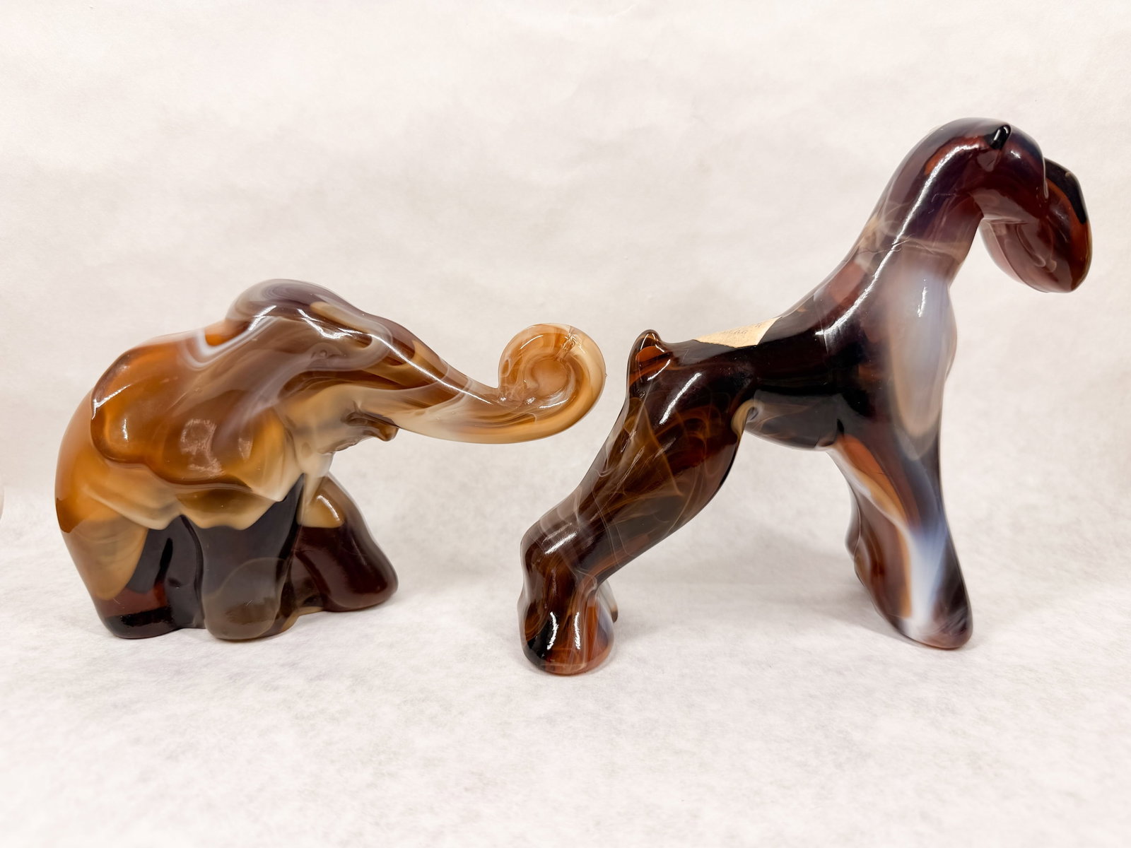 IMPERIAL CARAMEL SLAG FIGURINES, CIRCA 1960: A pair of caramel slag glass animal figurines manufactured by Imperial Glass Corporation, consisting of a trunk-up elephant and a terrier. The terrier features an adhesive paper label reading