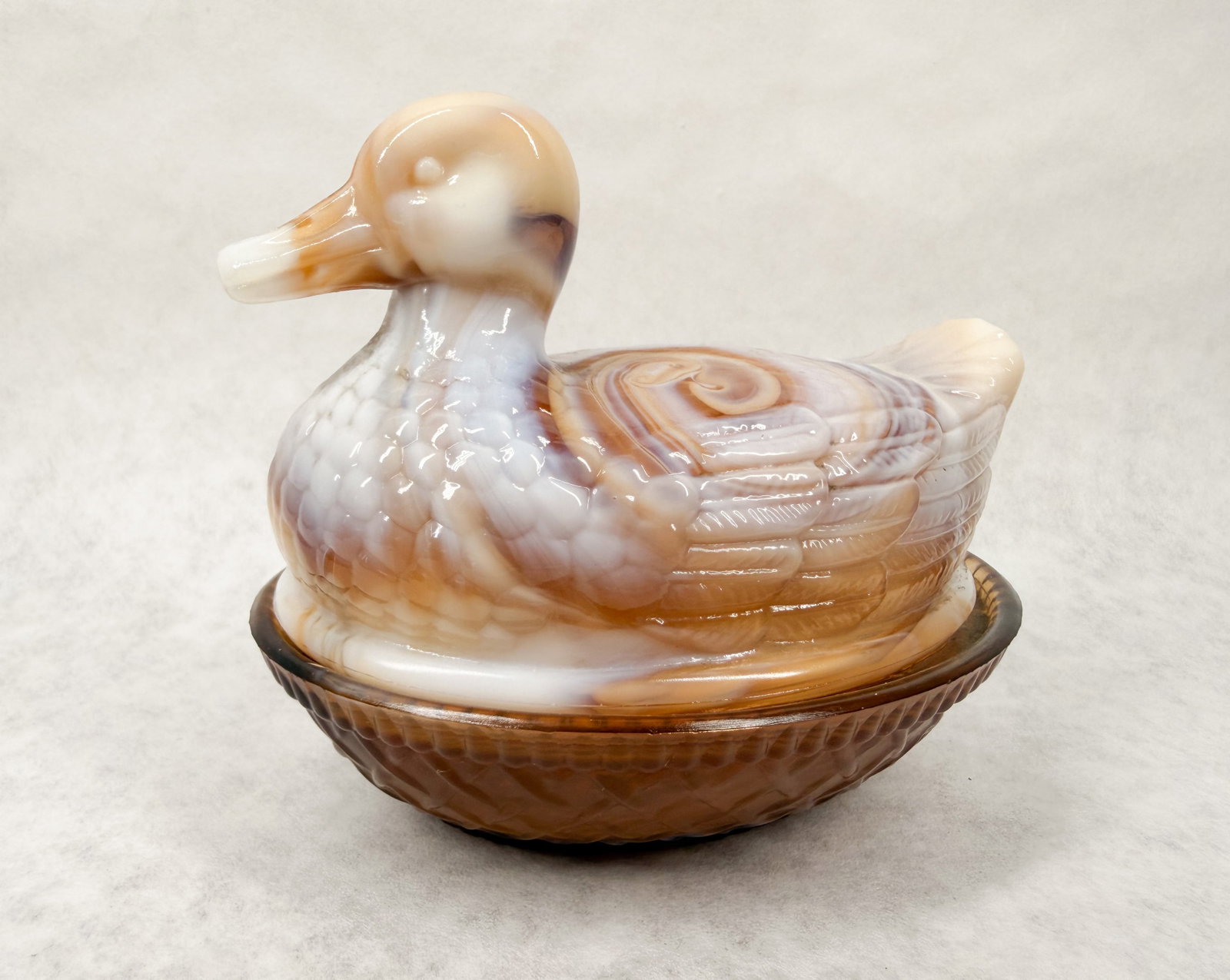 IMPERIAL CARAMEL SLAG GLASS DUCK DISH: A vintage slag glass covered dish modeled as a duck on a woven nest. An embossed monogram consisting of a capital letter G superimposed over a capital letter I is visible on the interior center of