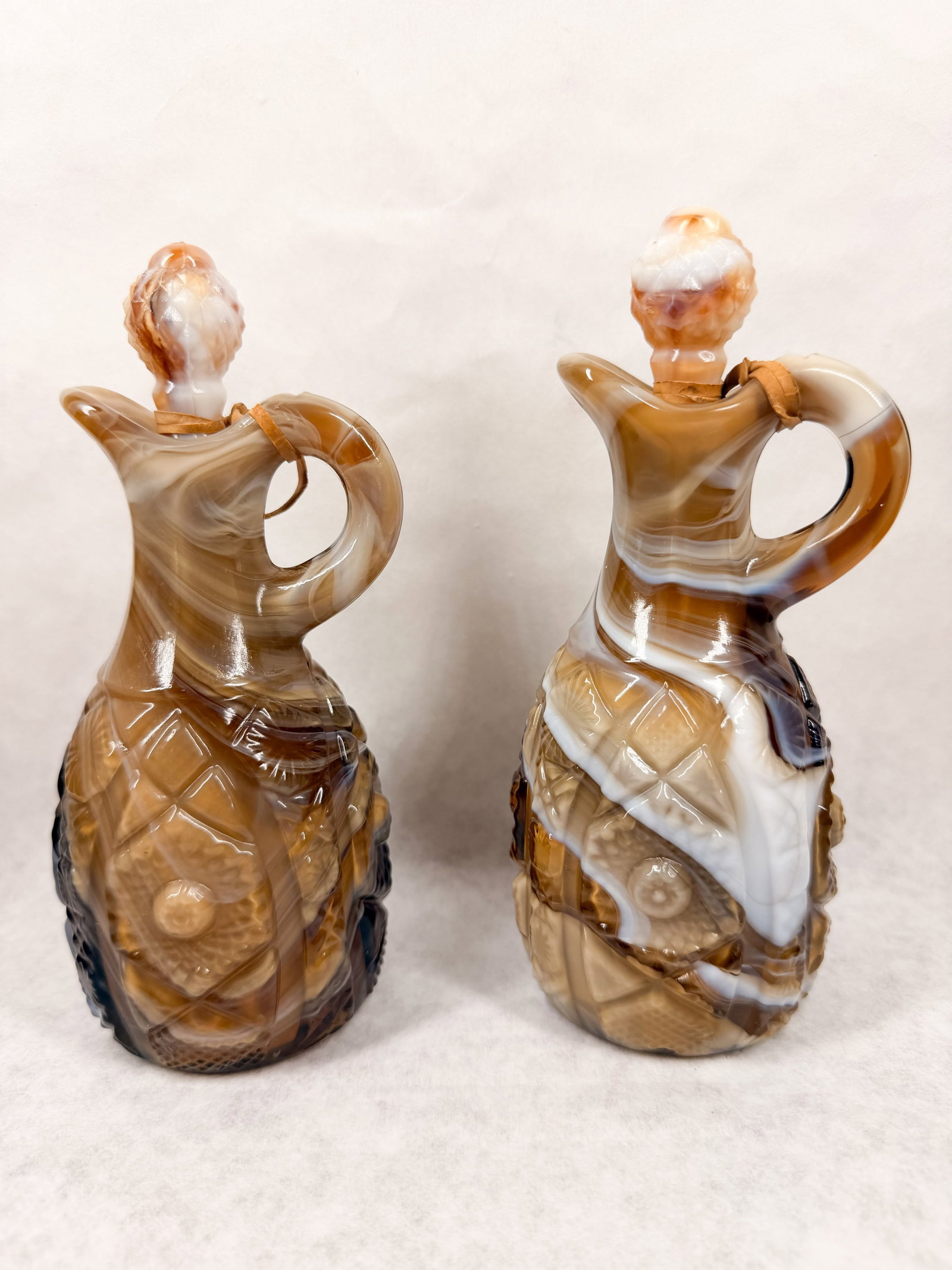 IMPERIAL PAIR OF CARAMEL SLAG GLASS CRUETS: A pair of pressed slag glass cruets in swirled tones of brown and cream. The decorative forms feature molded diamond and rosette motifs with matching pineapple-textured stoppers and