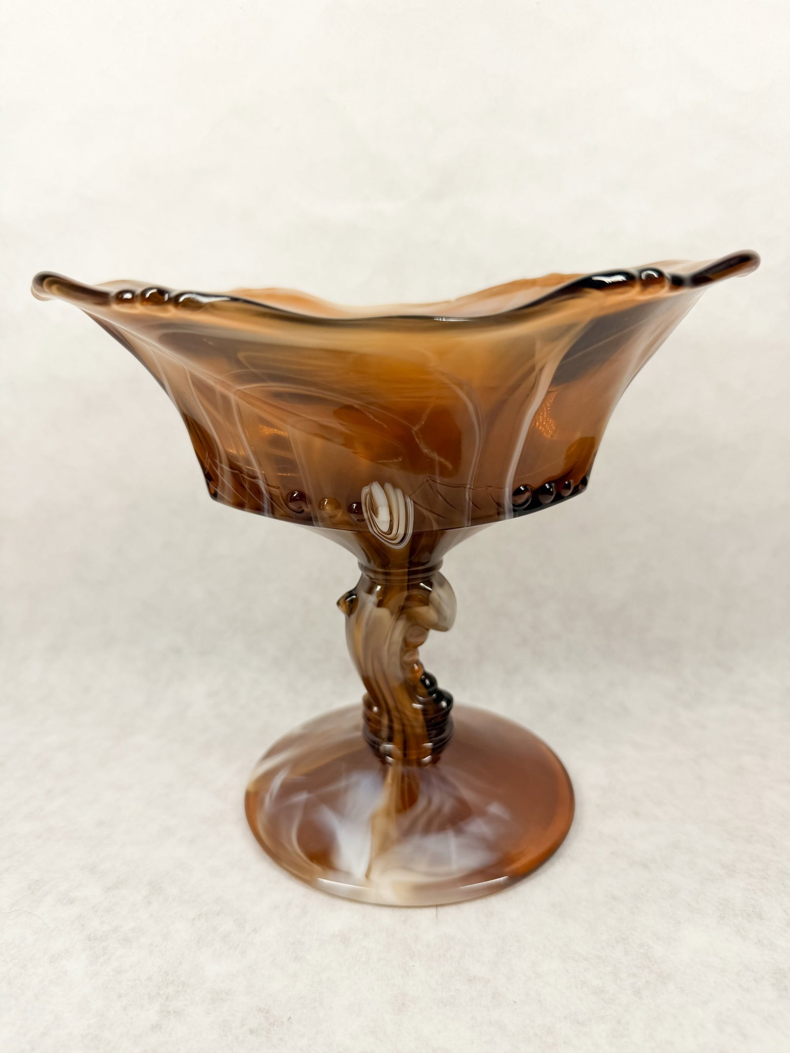 IMPERIAL CARAMEL SLAG COMPOTE: A molded slag glass compote featuring a ruffled bowl with a beaded band and caramel marbling. The vessel is supported by a sculptural pedestal base modeled as a stylized dolphin.